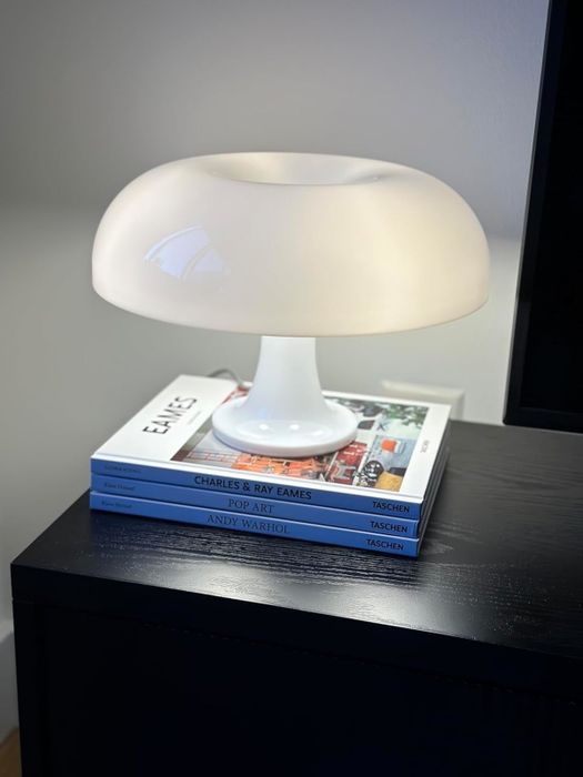 A sleek, white lamp placed on top of a stack of three art books: 'Charles & Ray Eames,' 'Pop Art,' and 'Andy Warhol.'
