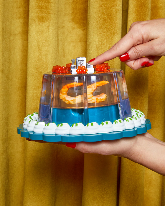 A hand holding a retro jelly mold ring lighter, decorated with shrimp and garnish.