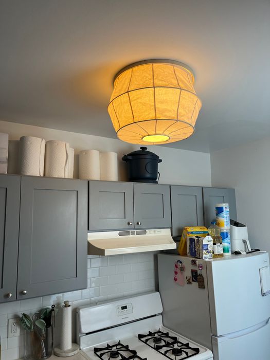 A kitchen with grey cabinets, a gas stove, paper towels, a rice cooker on top of cabinets, and a lit ceiling light. The fridge is adorned with magnets and small items placed on top