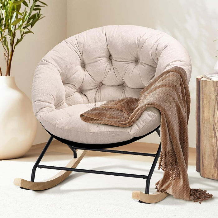 A round cushioned rocking chair, adorned with a soft, textured throw, placed in a cozy living space next to a wooden side table