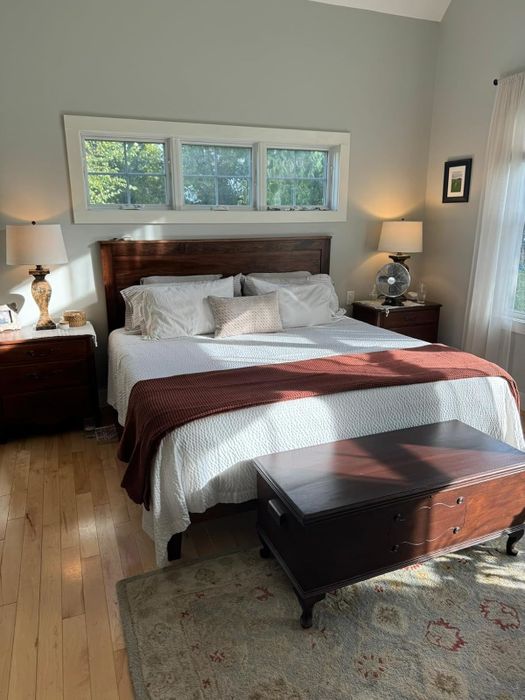 A well-lit bedroom with a wooden bed, white linens, a brown throw blanket, bedside tables, and a decorative chest at the foot of the bed.
