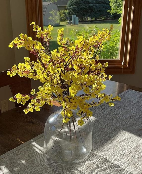 A sunny room with a glass vase on a table holding a bouquet of yellow flowers; a view of a lush green yard through the window in the background.