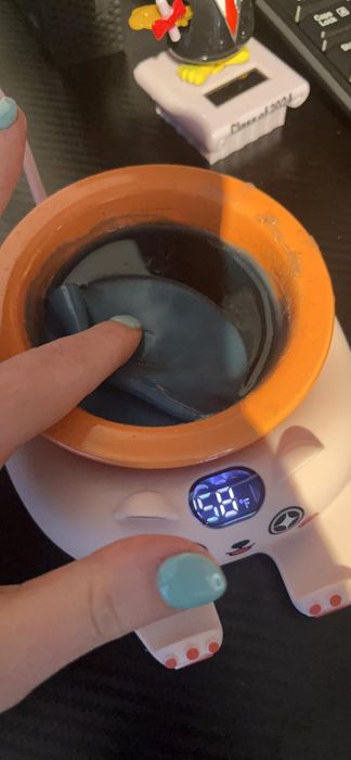 A hand dips a finger into a heated wax bath, showing 68°F, on a white cat-shaped melter.