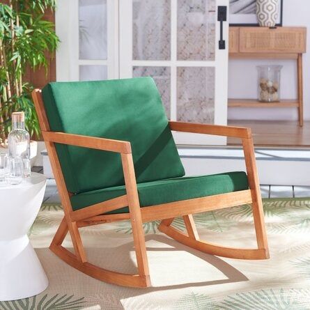 A wooden rocking chair with green cushions placed on a patio, next to a side table adorned with a glass decanter and glasses