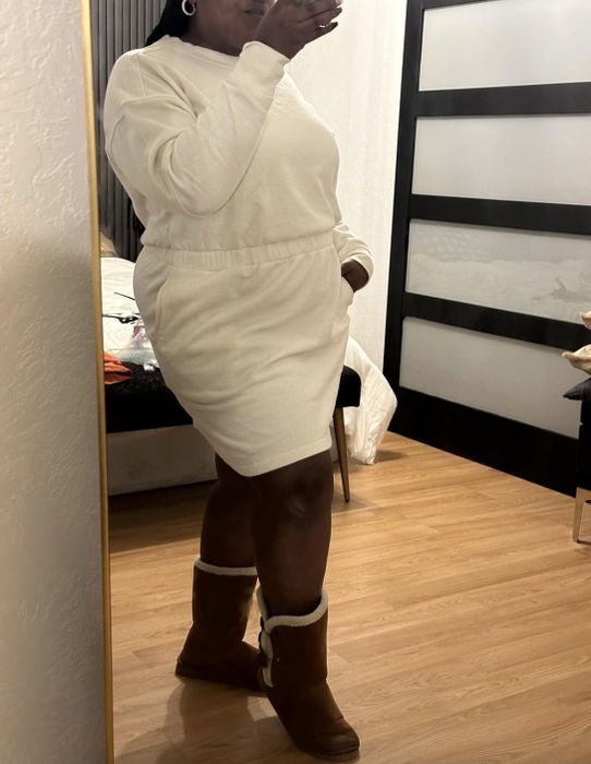 A reviewer takes a mirror selfie wearing a cozy, chic look consisting of a white sweater dress and brown boots.
