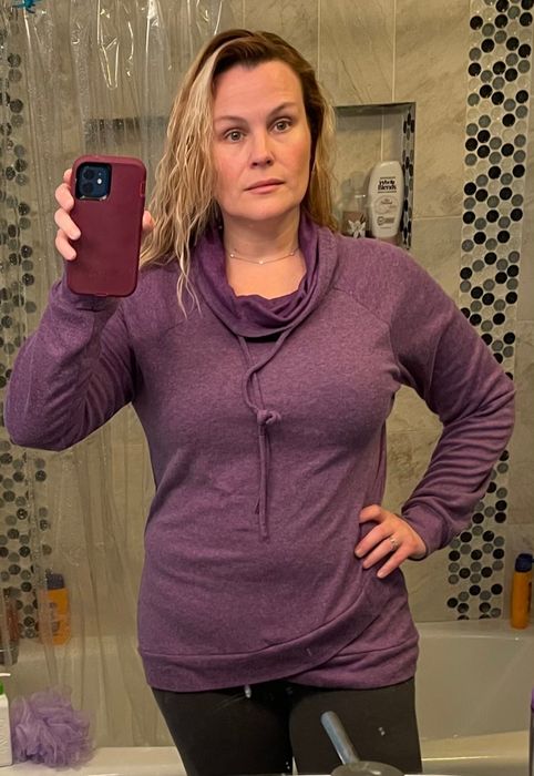 A person snaps a selfie in a bathroom mirror, wearing a casual purple long-sleeve top paired with dark pants, exuding a relaxed vibe.