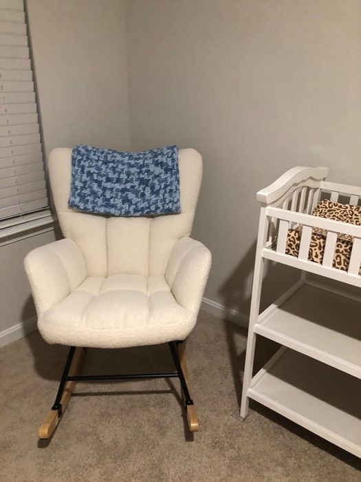 A warm white rocking chair with a blue knitted blanket draped over the back, beside a white changing table with a leopard-print cover in the corner of a nursery