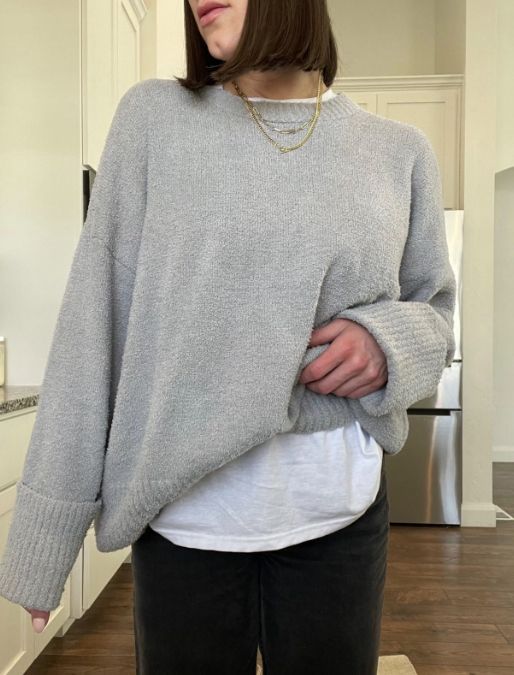 Reviewer posing indoors, wearing a cozy grey oversized sweater with a white shirt underneath, dark pants, and accessorized with a layered gold necklace.