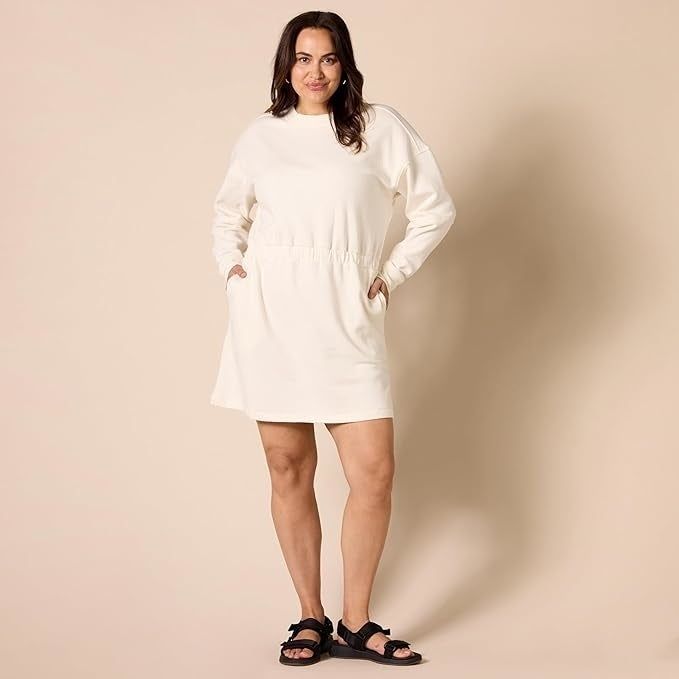 A model showcases a laid-back, long-sleeve dress with pockets, paired with black sandals, standing against a simple backdrop.