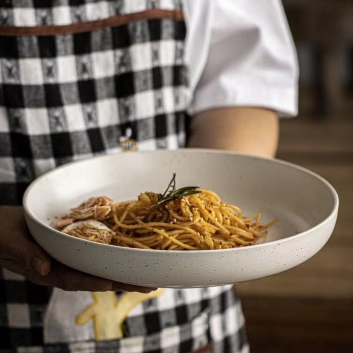 A person wearing a checkered apron holding a white plate of spaghetti, topped with shrimp, showcasing a delicious food dish.