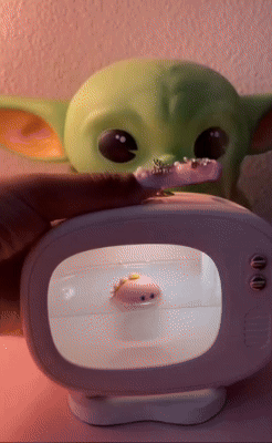 A miniature Baby Yoda toy sits in the background with a cute pink toy displayed inside a small TV-shaped display case in the foreground.