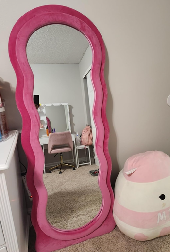 A full-length mirror with a wavy pink frame, placed next to a white dresser and a cozy plush chair, contributing to a chic room decor setup.