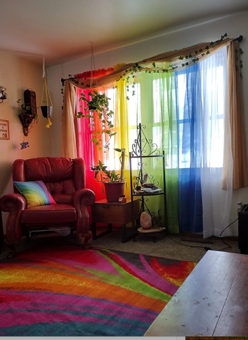 A cozy living room featuring colorful sheer curtains, a red armchair, plants, and a vibrant rug with swirling patterns.