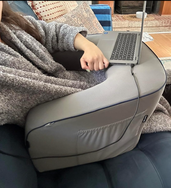 A person using a laptop while seated on a couch, with the laptop resting on a cushioned lap desk. The setting conveys comfort and convenience for home work or casual browsing