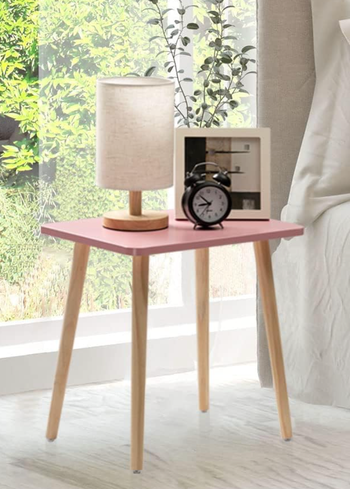 A compact side table with a lamp, a clock, and a framed photo beside a bed, positioned next to a window where greenery peeks outside.