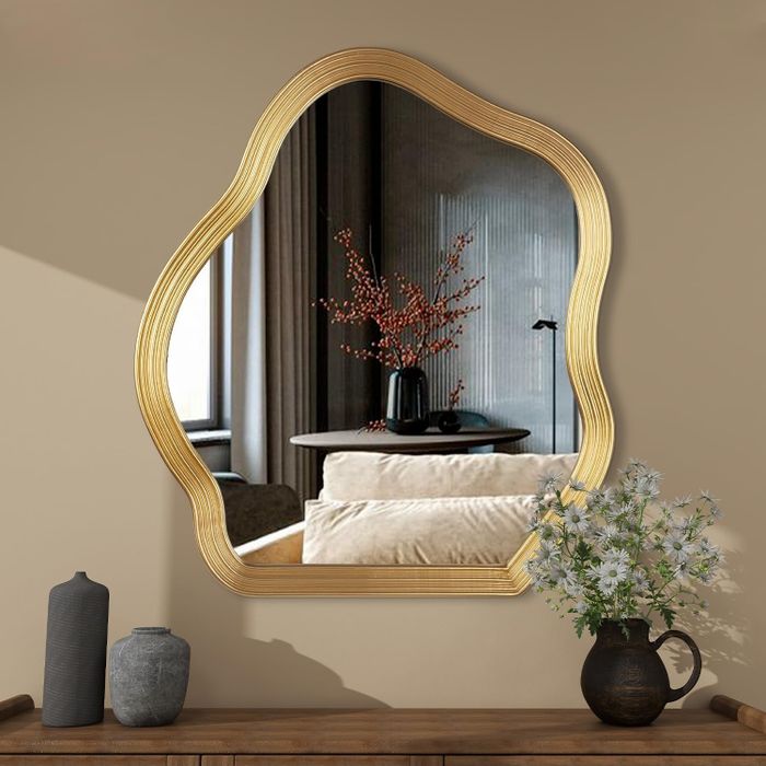 A decorative mirror with a wavy gold frame reflecting a cozy living room scene, complete with a sofa, side table, black vases, and orange berry branches in a vase.