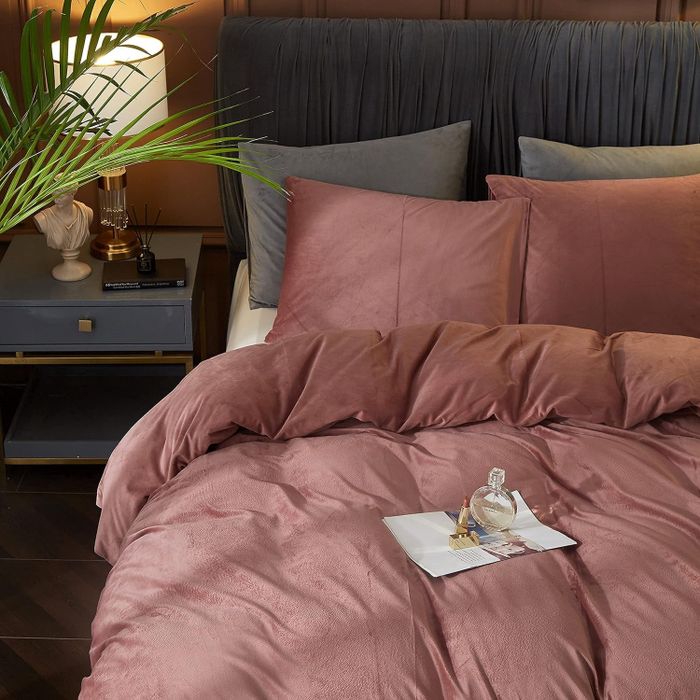 A lavish bedroom setup featuring a plush duvet, coordinating pillows, a nightstand with a stylish lamp and a plant, and a tray holding perfume and a book.