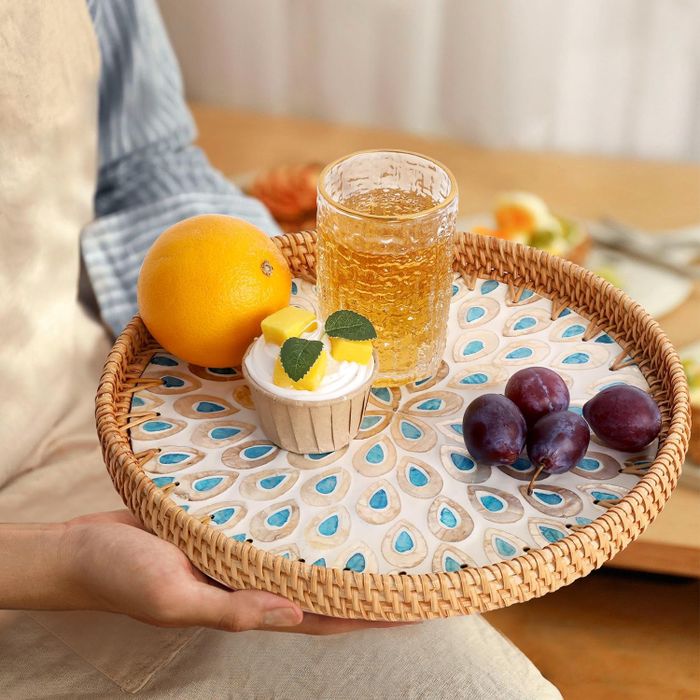 A woven tray displaying a glass of juice, an orange, a frosted cupcake, and three plums, creating a stylish and attractive setting for kitchenware shopping.