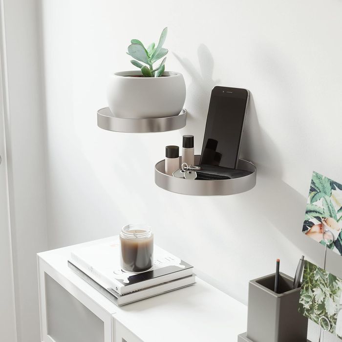 Two floating wall shelves displaying a potted plant, a smartphone, lip balm, and keys. Below, a console table holds a candle, some closed books, and a pen holder.