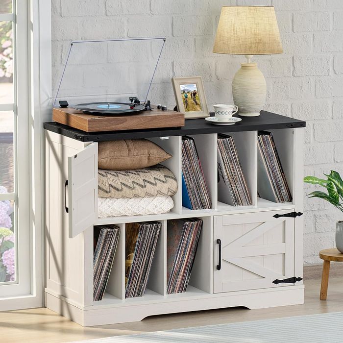 A record player resting on a cabinet filled with vinyl records and stacked blankets, topped with a lamp and framed photo. A cozy, modern living room vibe