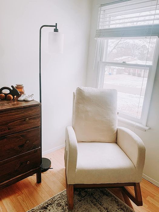 A reviewer photo of a white rocking chair placed in the corner of a room near the window.