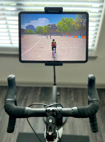 A stationary bike setup with a tablet attached, displaying a virtual cycling simulation with cyclists riding along a paved path in a park setting