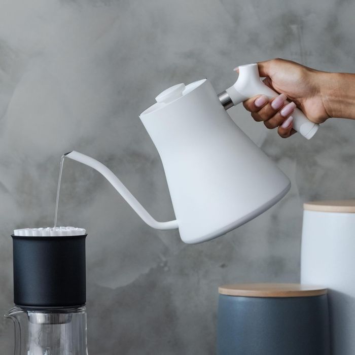 A hand gracefully pours hot water from a white gooseneck kettle into a black filter positioned over a glass container.