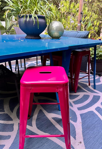A pink metal bar stool placed at a blue outdoor table, accompanied by a potted plant and a green decorative sphere. Rustic chic styling perfect for patio shopping inspiration.