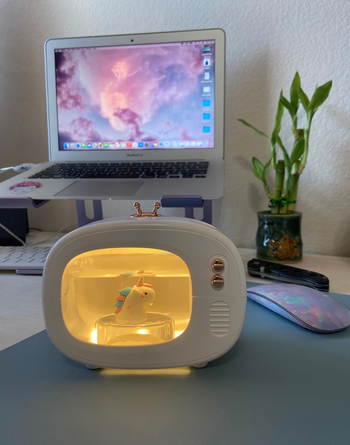 A charming unicorn lamp, nestled in a retro-style TV casing on a desk, complete with a computer and bamboo plant in the background.
