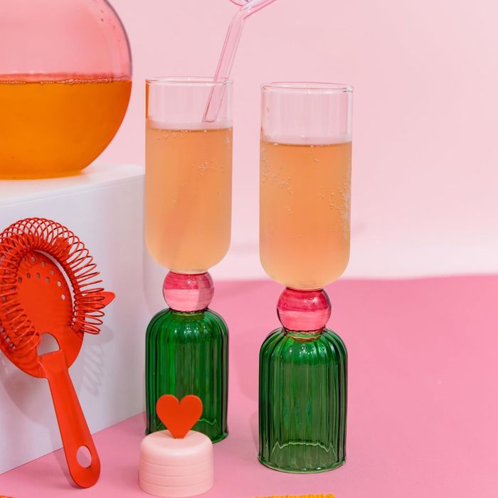 Two contemporary champagne flutes with vibrant bases, filled with bubbly, alongside a festive drink stirrer and a heart-shaped bottle stopper.