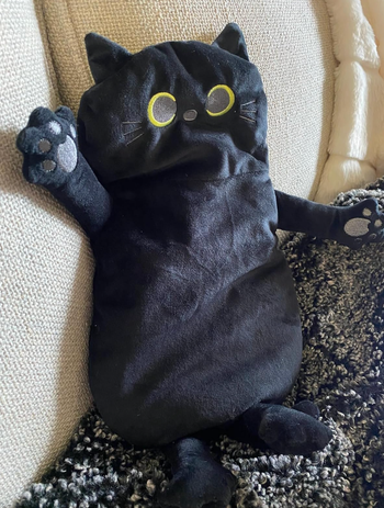 A plush black cat pillow with bright yellow eyes, resting on a couch, perfect for home decor or as a snuggly gift.
