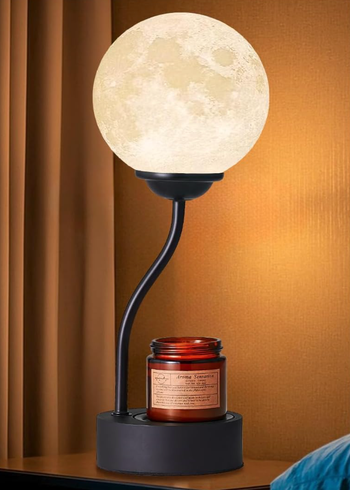 A moon-shaped lamp with a curved stand sits beside a red candle on a modern side table, radiating a warm, cozy atmosphere.