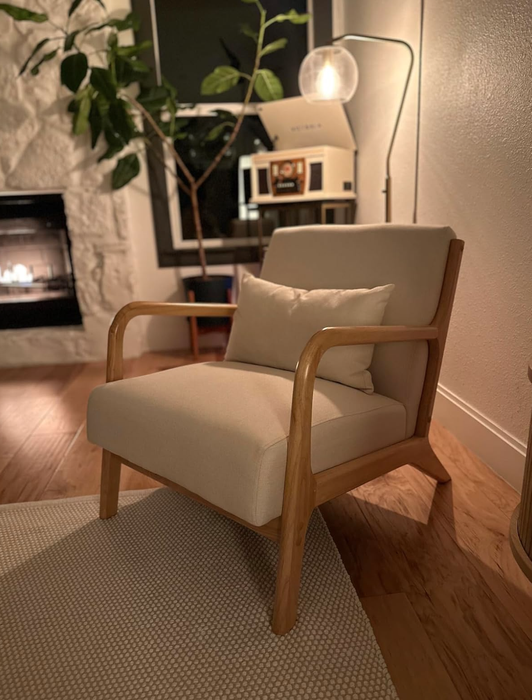 A modern living room featuring a stylish wooden armchair with beige cushions, illuminated by a floor lamp next to a window with a plant.