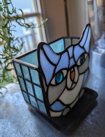 A beautifully detailed stained glass owl candle holder resting on a windowsill beside a lush plant.