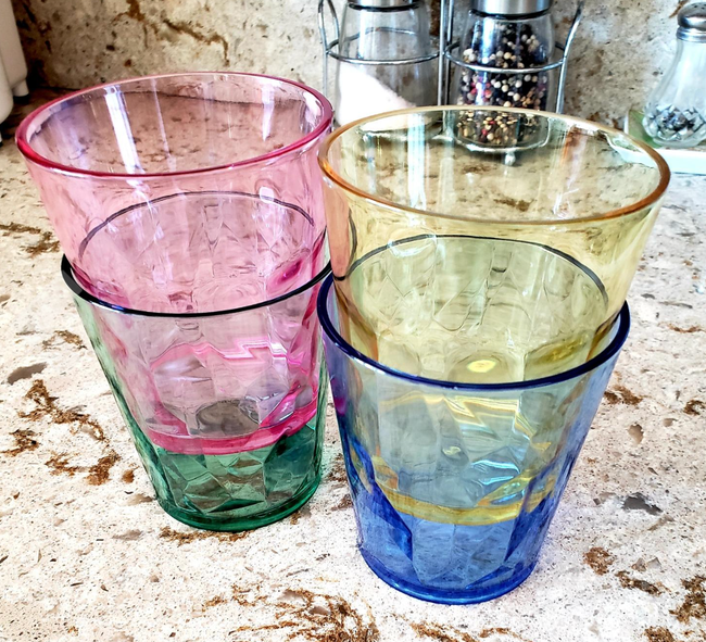Four textured drinking glasses, each in a different hue, neatly stacked on a kitchen counter with various kitchen items in the background.