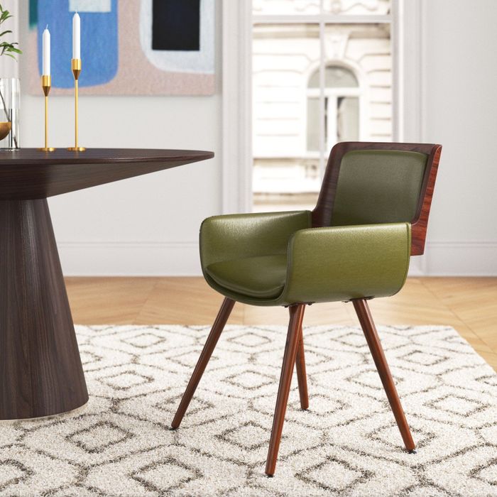 A modern green cushioned chair with wooden legs positioned on a patterned rug, beside a dark wooden table adorned with candlesticks, and near a window.