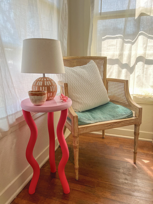A chic seating area with a woven chair, teal cushion, and a wavy-legged pink table. The table holds a ceramic lamp and bowl, creating a cozy and inviting vignette.