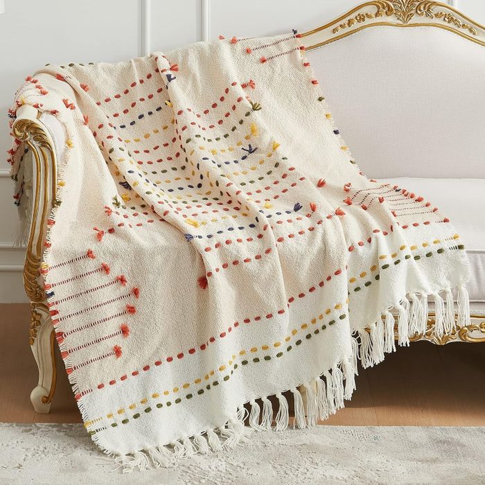 A white woven throw blanket with colorful dotted stripes and fringe edges draped over a gold-accented white sofa, creating a touch of warmth and style.