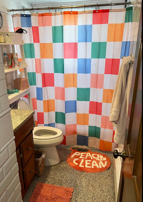 A bathroom featuring a multicolored checkered shower curtain, toilet, sink, and shelves stocked with toiletries.