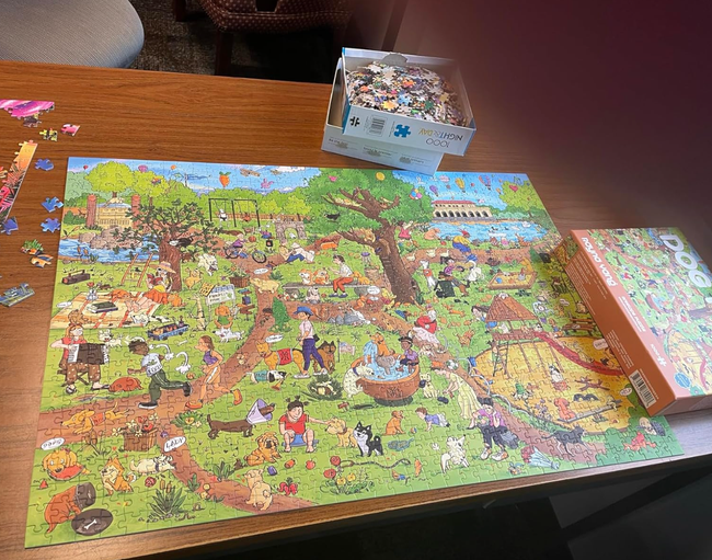 A completed puzzle of a bustling park scene rests on a table, alongside the puzzle box