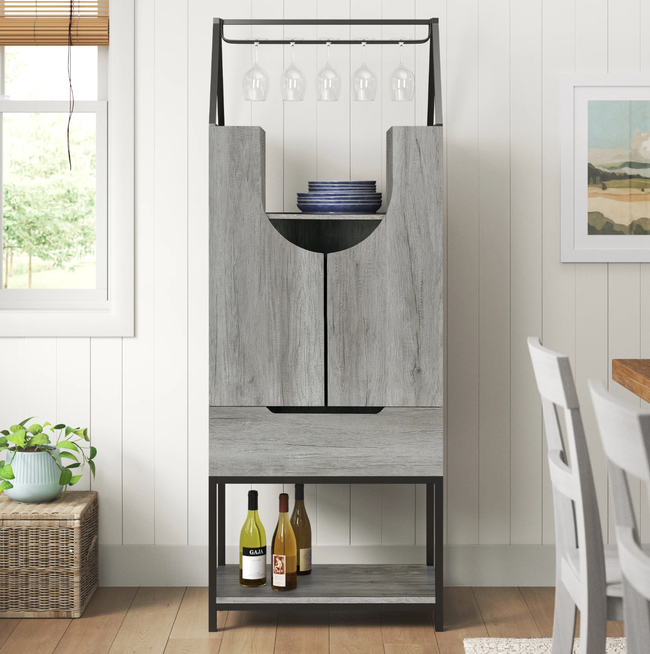 Tall gray wooden bar cabinet with bottles and glasses neatly arranged on top