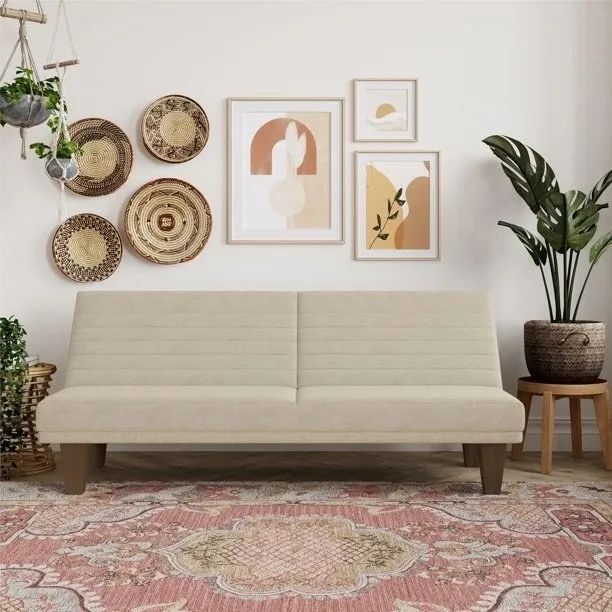 A tan futon placed in a cozy living room with a rug and artwork on the wall.