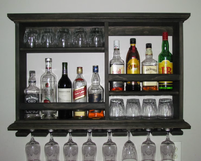 Small brown wooden bar cabinet mounted on the wall with bottles and glasses neatly arranged on shelves