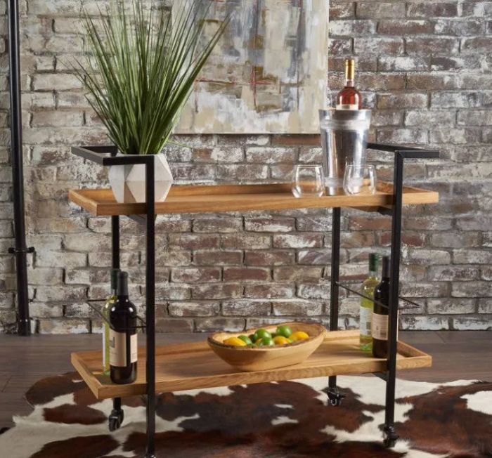 A two-tier bar cart with a black frame and light brown wooden shelves, sitting elegantly on a cowhide rug.