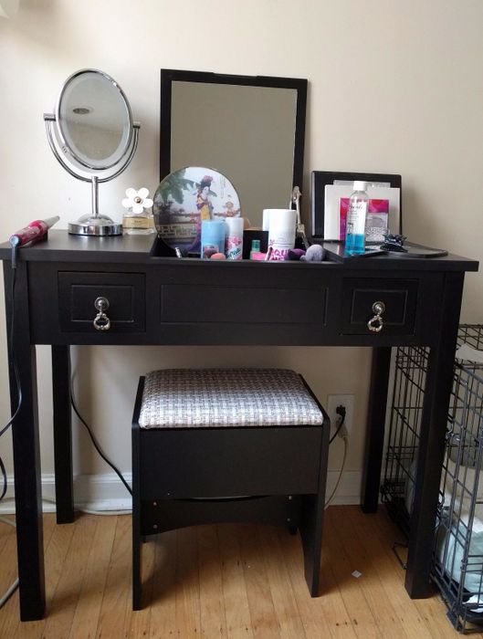 A reviewer's photo featuring a sleek black vanity table with a flip-up mirror, products neatly arranged on top, and a stool tucked underneath