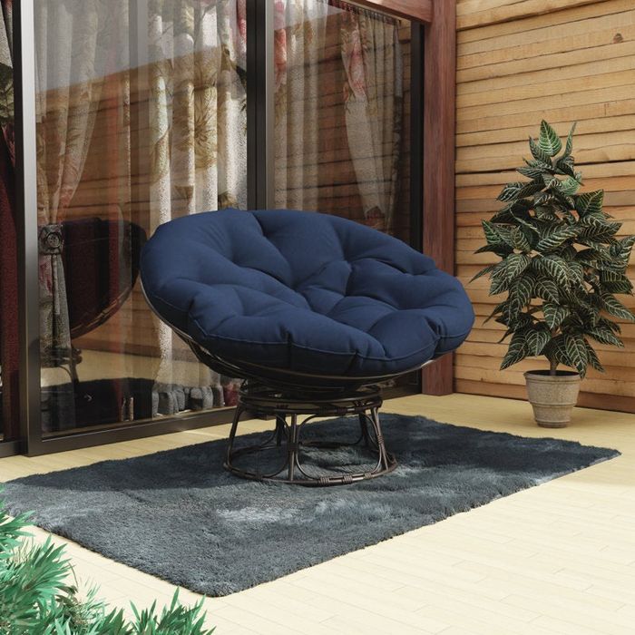 A cozy Papasan chair with a plush, tufted cushion sitting on a deck, beside a potted plant and large glass windows