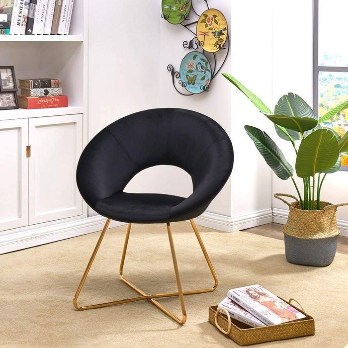 A sleek black chair with gold metal legs is placed in a chic living room, complemented by a plant and a basket of magazines for a cozy atmosphere