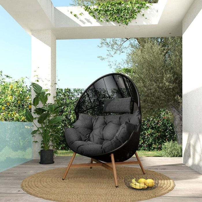 A cozy outdoor seating area featuring a round wicker chair, soft cushions, and a small bowl filled with lemons on a woven rug, surrounded by lush greenery.
