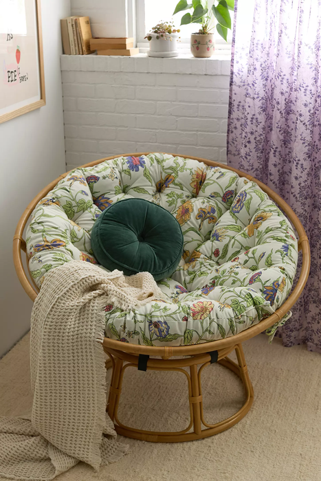 A papasan chair with a floral cushion and a green throw pillow, placed next to a cozy cream knit blanket in a corner with purple curtains and a potted plant, creating a serene nook.
