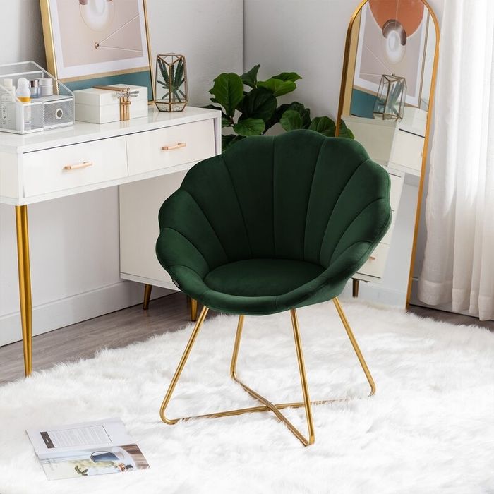 A chic green scalloped velvet chair with gold legs, placed in a vanity corner, complemented by a white fur rug, a potted plant, and framed mirrors that enhance the room’s stylish vibe.